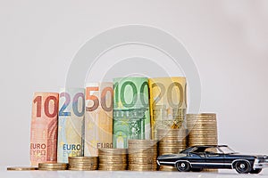 A black model of a car and ten, twenty, fifty, one hundred, two hundred and coins euro rolled bills banknotes on white background