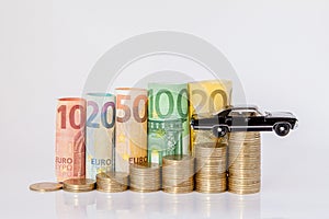 A black model of a car and ten, twenty, fifty, one hundred, two hundred and coins euro rolled bills banknotes on white background
