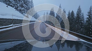 Black modern car moves on road in rural area along snow covered trees and hills.