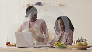 Black mother teaching her little daughter to cook vegetable salad, girl cutting vegetables at kitchen