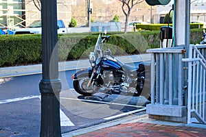 A black motorcycle parked on the street surrounded by a staircase, lush green plants, a red brick sidewalk and bare winter trees