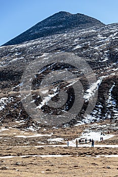 Black mountain witn snow and below with tourists on the ground with brown grass, snow and frozen pond in winter at Zero Point.