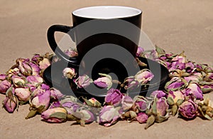 A black mug with a saucer with tea made from dried buds and petals of a purple damask rose stands on a paper background