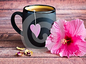Black mug of tea with a pink heart on a string Pink hibiscus flower on dark wood background