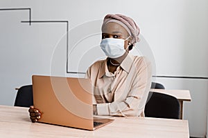 Black muslim business woman in medical mask with laptop. Working on line at home. Cryptocurrency investments in bitcoin