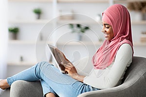 Black Muslim Female In Hijab Relaxing With Digital Tablet At Home