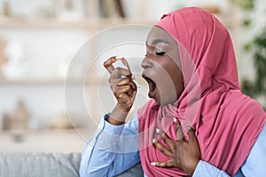 Black muslim woman in hijab using inhaler at home, suffering breathing problem