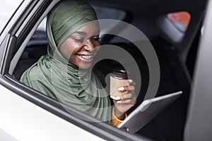 Black muslim woman riding car on backseat with digital tablet and coffee