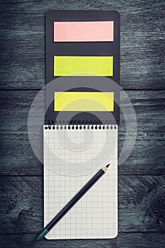 Black notebook with a sheet in a cage with pencil and colored sticky notes on a wooden rustic background, top view