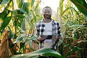 Black notepad is in hands. Young African American man is standing in the cornfield at daytime