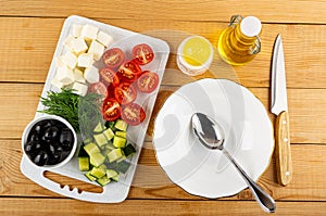Black olives, feta cheese, dill, pieces of cucumbers, tomato on cutting board, spoon in bowl, bottle of vegetable oil, salt, knife
