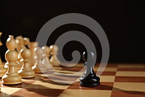 Black pawn in front of white pieces on wooden board against dark background. Competition concept