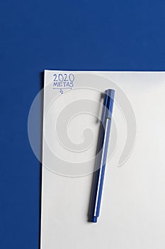 Black pen with blue on classic blue desk with paper sheet ready for 2020 goals in Spanish written in blue ink