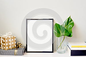 Black photo frame, green plant in a crystal vase, gift boxes and a pile of books arranged against empty grey wall. Frame mock-up.