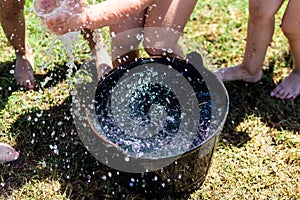 Black plastic bucket with fresh water with many kids legs playing summer games on a green grass meadow