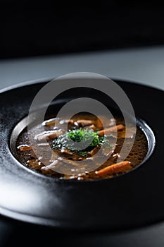 black plate with brown soup pottage with mushroom, carrot, vegetables, dill