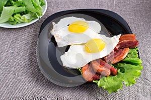 Black plate with fried eggs, bacon and lettuce closeup