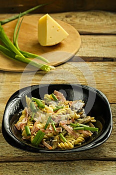 Black plate with pasta with ham and mushrooms in a creamy sauce on the table with a fork on the board.