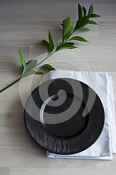 Black plate on white wooden background. With plant and napkin,top view. Empty plate for serving