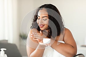 Black plus size lady applying body cream on shoulder after shower in morning, making beauty procedure