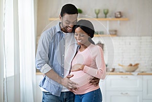 Black pregnant woman and her handsome husband embracing and smiling while spending time together near window at home