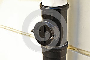 black PVC downpipe rain water leader or gutter closeup. round bolted cleanout plate. white stucco exterior wall