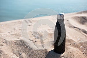 Black reusable steel stainless thermo water bottle in sand on the beach.