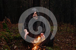 A black-robed monk sits by a fire in the forest and looks into a divination ball. Mysticism and magic, sorcerers and witches 3