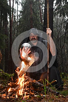 A black-robed monk sits by a fire in the forest and looks into a divination ball. Mysticism and magic, sorcerers and witches 3