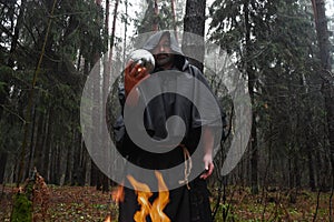 A black-robed monk sits by a fire in the forest and looks into a divination ball. Mysticism and magic, sorcerers and witches 3