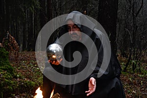 A black-robed monk sits by a fire in the forest and looks into a divination ball. Mysticism and magic, sorcerers and witches 3