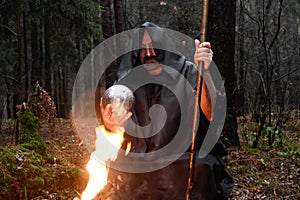 A black-robed monk sits by a fire in the forest and looks into a divination ball. Mysticism and magic, sorcerers and witches 3