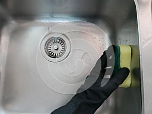 Black rubber gloved hand holding a sponge and cleaning a stainless steel sink