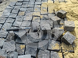 A black rubber mallet lies in the midst of natural gray granite stones and yellow sand. Cobblestone paving - tools and materials