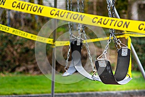 Black rubber swings in closed public playground surrounded by yellow caution tape during Corvid-19 Coronavirus pandemic