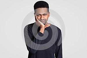 Black sad young man looks with dejected expression, holds chin, has short haircut, dark skin, feels unhappy and lonely, wears