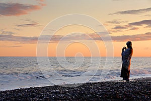 Black-sand beach at sunset. Young woman taking pictures with her mobile phone.