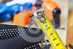 The black screw in hand and is measured by a yellow tape measure with a ruler on a wooden board background. The concept of tools