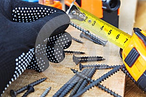 The black screw in hand and is measured by a yellow tape measure with a ruler on a wooden board background. The concept of tools