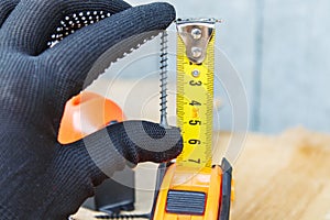 The black screw in hand and is measured by a yellow tape measure with a ruler on a wooden board background. The concept of tools