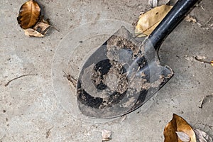 A black shovel with dirt caked on it from digging lays on a sandy concrete surface with brown winter leaves scattered around