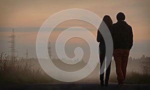 Black silhouette of couple walking along misty street in fog after sunset. View from behind. Man holding woman