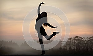 Black silhouette of pregnant woman standing on one leg. Other foot and hands raised in air. Foggy evening landscape