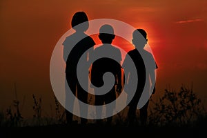 Black silhouette of three children standing together. There is a sky at sunset