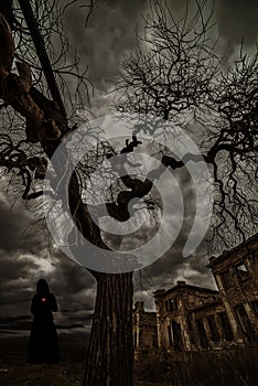 Black silhouette of a woman in a hoodie and terrible trees with branches against a background of terrible ruins.