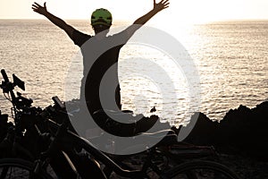 Black silhouette of woman with yellow helmet in front to the sea under sunset light. Arms raised. Freedom and happiness concept. A photo