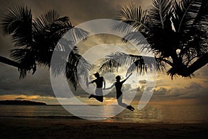 Black silhouette of young couple jumping in the air on tropical beach during sunset on background of palm trees and sea