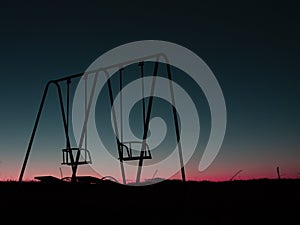 Black silhouettes of a couple of metal abandoned empty swings at dusk