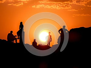 Black silhouettes of people on a rock against an orange sky looking at a beautiful, setting sun