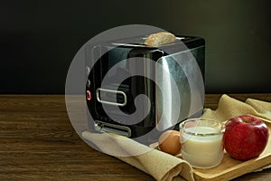 Black and silver electric micro oven with toast, fresh eggs, red apple and glass of milk on brown wooden and black background,